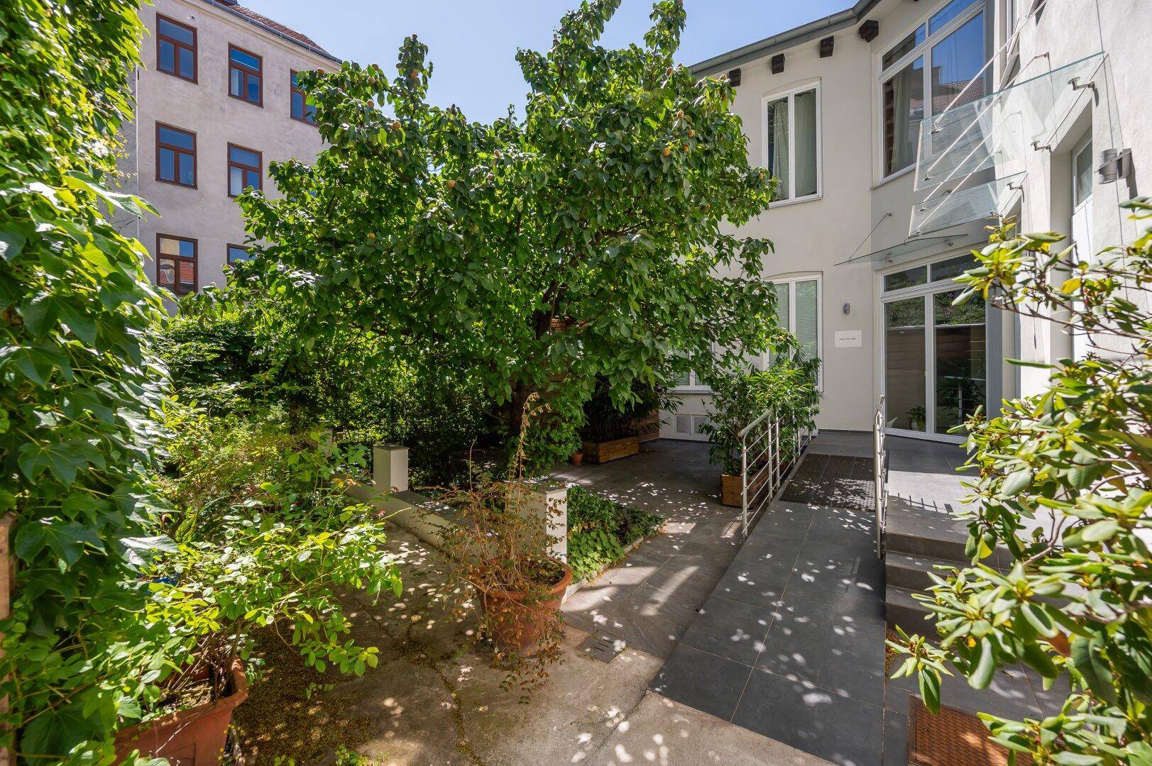 Grüner Innenhof mit Bäumen, Pflanzen und einer barrierefreien Rampe zum Eingang.