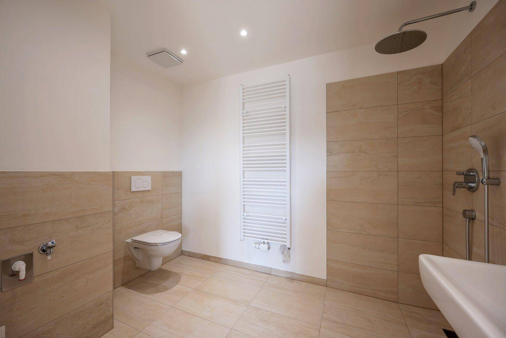 Modernes Badezimmer mit beigefarbenen Fliesen, wandhängendem WC und großer Regendusche.