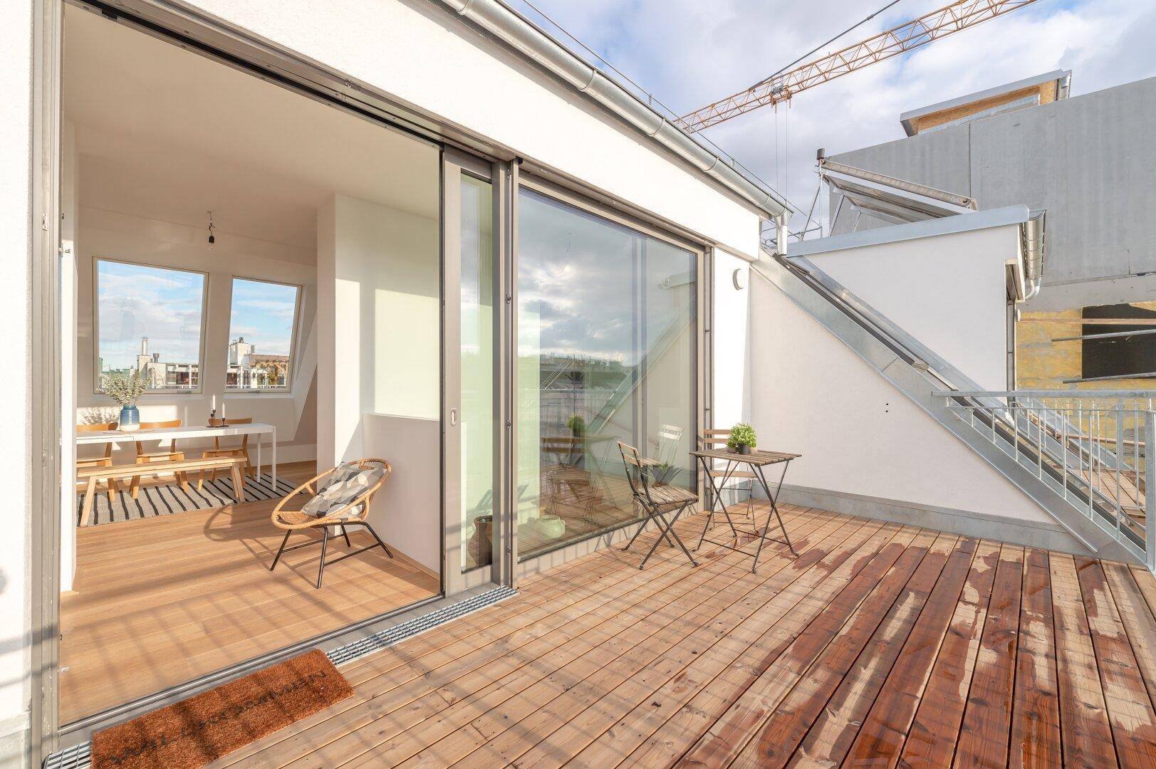 Sonniger Balkon mit Holzboden und Blick auf die Umgebung, verbunden mit dem Innenbereich durch eine Schiebetür.