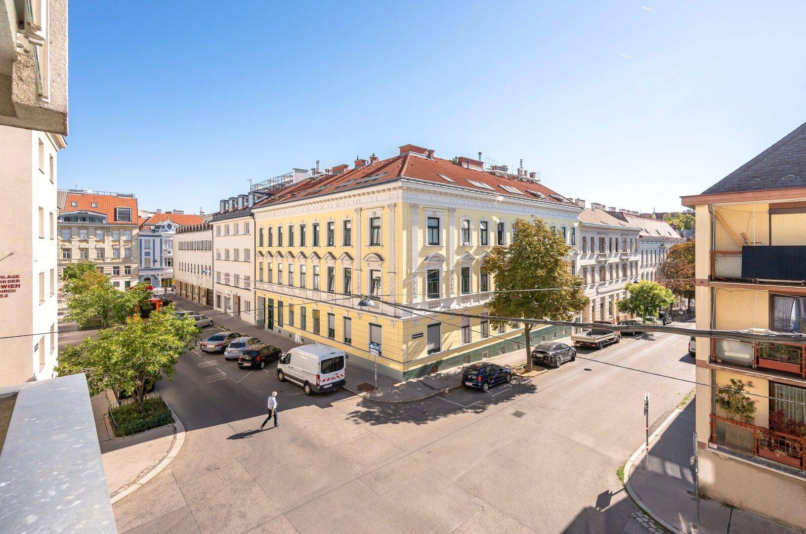Weitläufiger Blick über eine belebte Stadtstraße mit historischen Gebäuden und viel Grün.