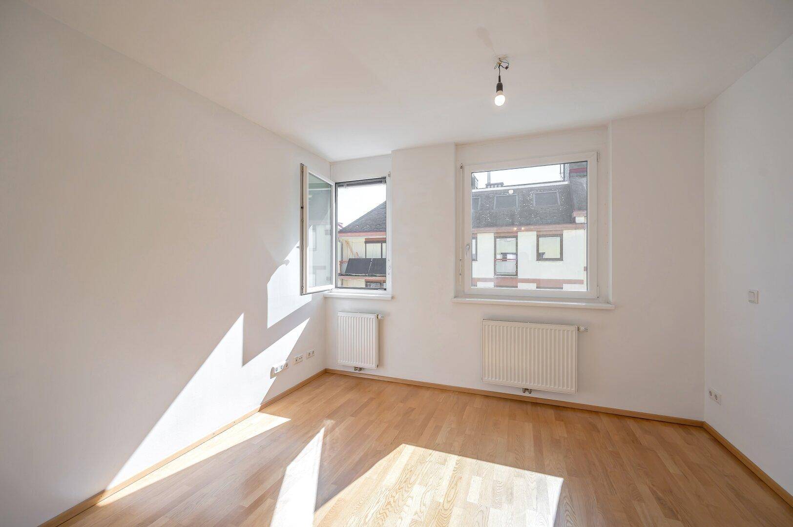 Helles Schlafzimmer mit Parkettboden und zwei Fenstern, die viel Tageslicht hereinlassen.