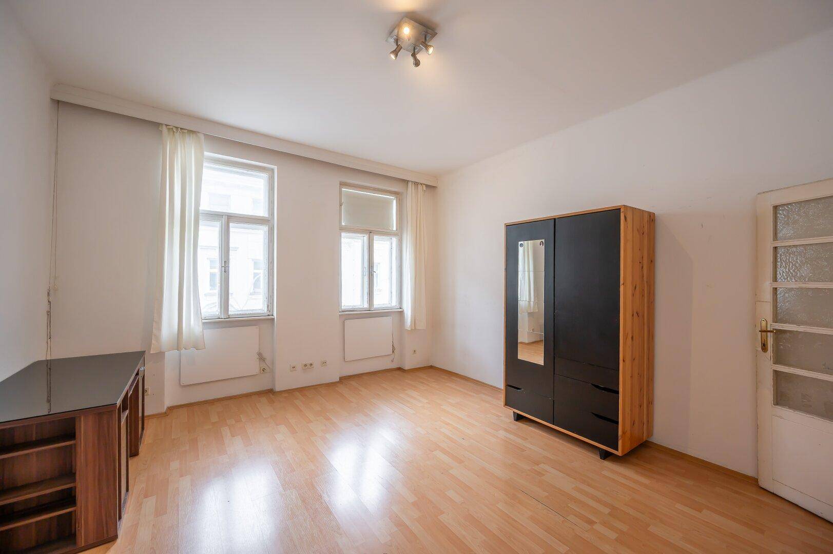 Helles, leeres Zimmer mit Holzboden, zwei Fenstern und einem Kleiderschrank.