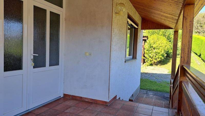 Überdachter Eingangsbereich mit weißer Tür und Fliesenboden, umgeben von Grün.