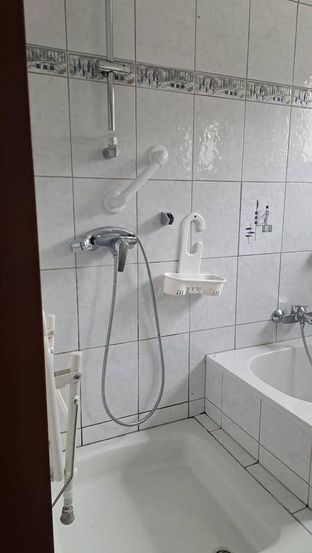 Badezimmer mit Badewanne, Duscharmatur und Wandfliesen in hellem Design, ausgestattet mit Haltegriffen.