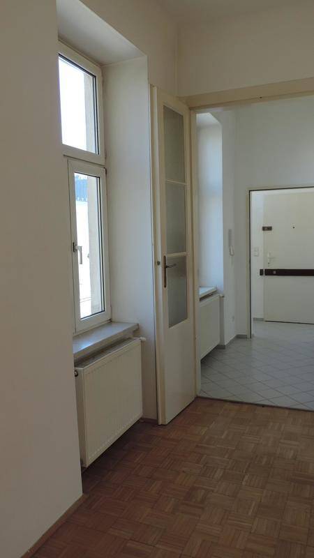 Heller Raum mit Parkettboden, Fenster und einer Tür, die zu einem gefliesten Bereich führt.