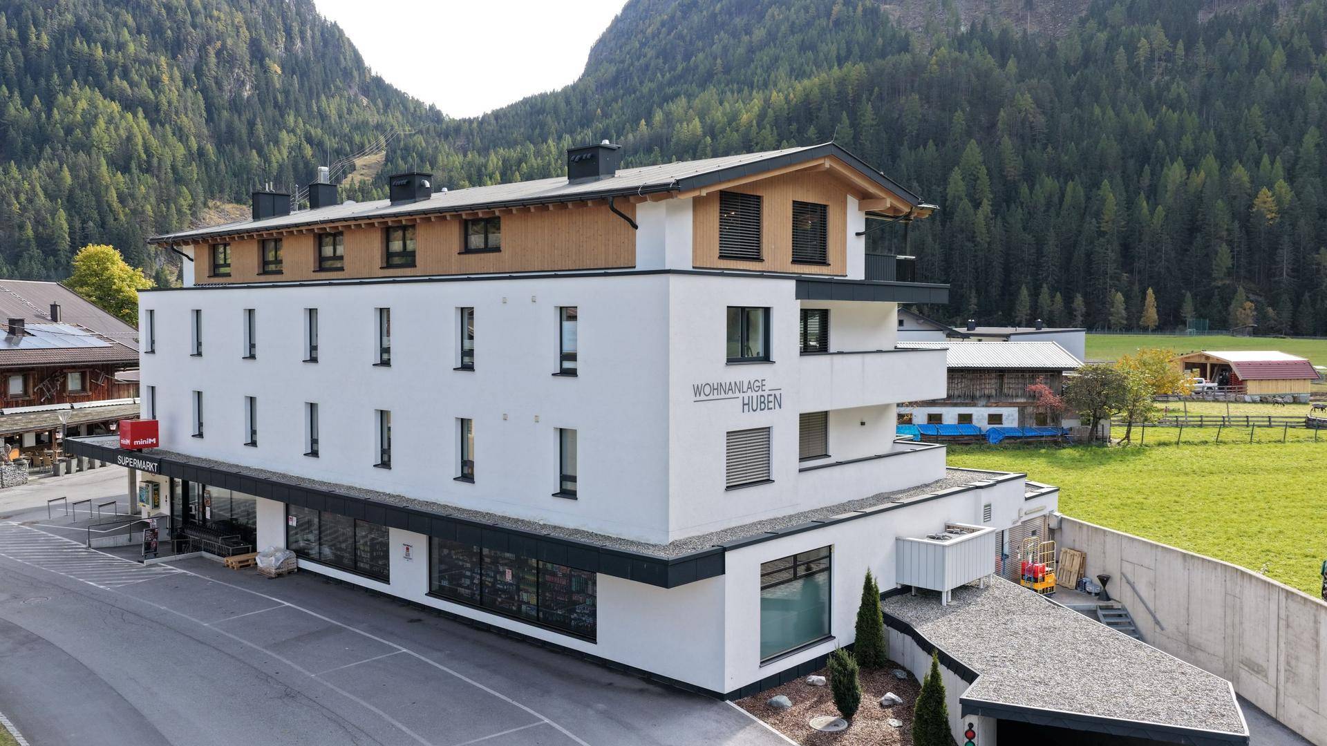Modernes Mehrfamilienhaus mit Supermarkt im Erdgeschoss, umgeben von alpiner Landschaft und Wäldern.