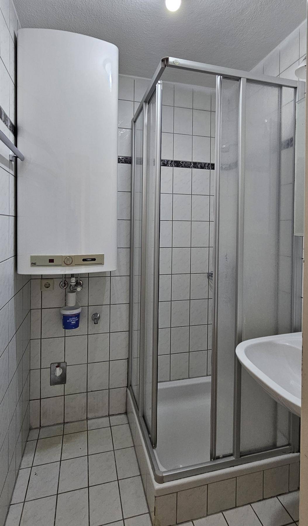 Kleines Badezimmer mit Duschkabine, Waschbecken und einem Warmwasserboiler, funktional eingerichtet.