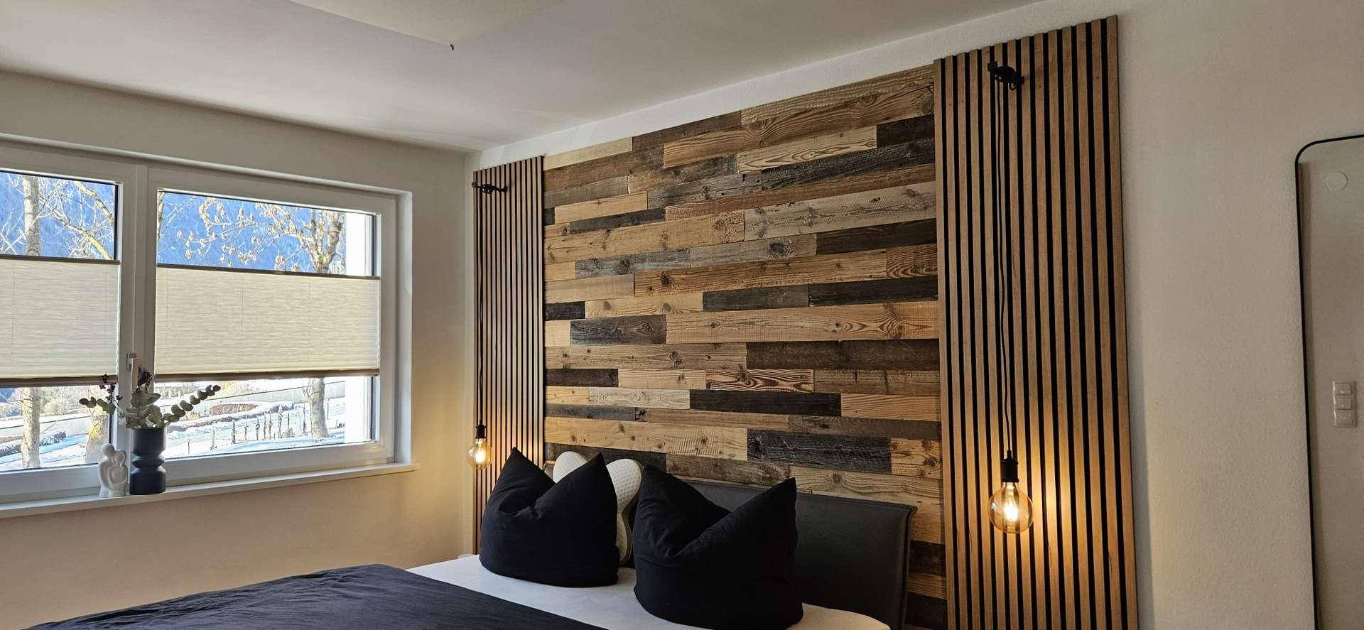 Schlafzimmer mit dekorativer Holzwandverkleidung, modernen Leuchten und großem Fenster mit Ausblick.