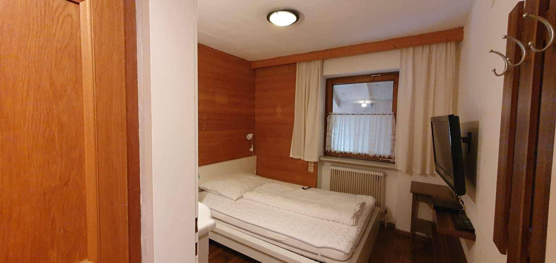 Kleines Schlafzimmer mit Einzelbett, Holzwänden und Fernseher an der Wand, hell durch das Fenster.