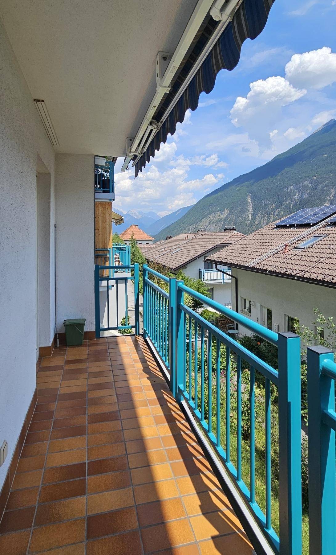 Sonniger Balkon mit blauer Brüstung und Fliesenboden, bietet einen weiten Blick auf die umliegenden Berge.