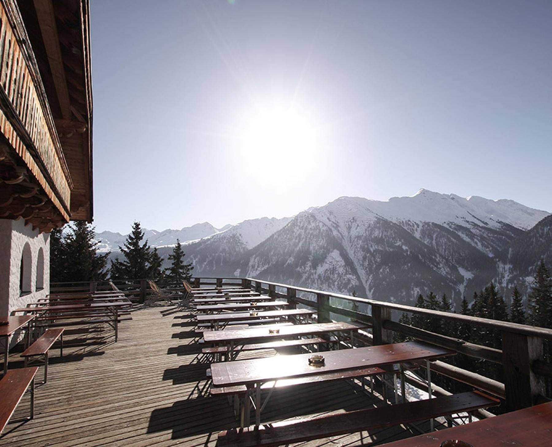 Sonnige Holzterrasse mit zahlreichen Sitzgelegenheiten und atemberaubendem Panoramablick auf die verschneiten Berge.