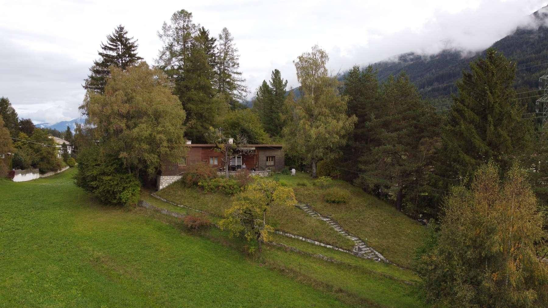 Luftaufnahme eines Hauses auf einem bewaldeten Hügel mit Bergen im Hintergrund unter bewölktem Himmel.
