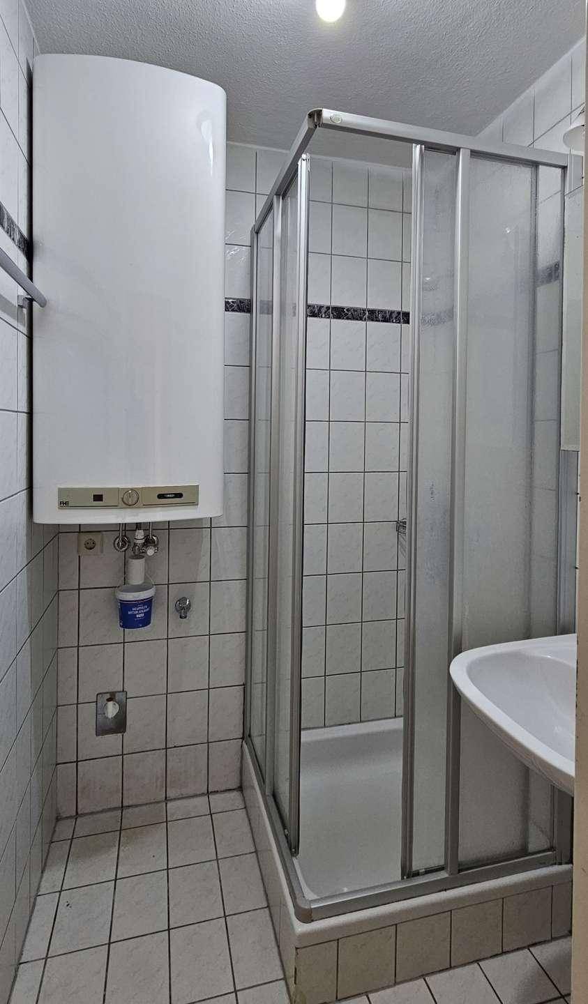 Kleines Badezimmer mit Duschkabine, Waschbecken und Wandboiler, komplett gefliest für einfache Pflege.
