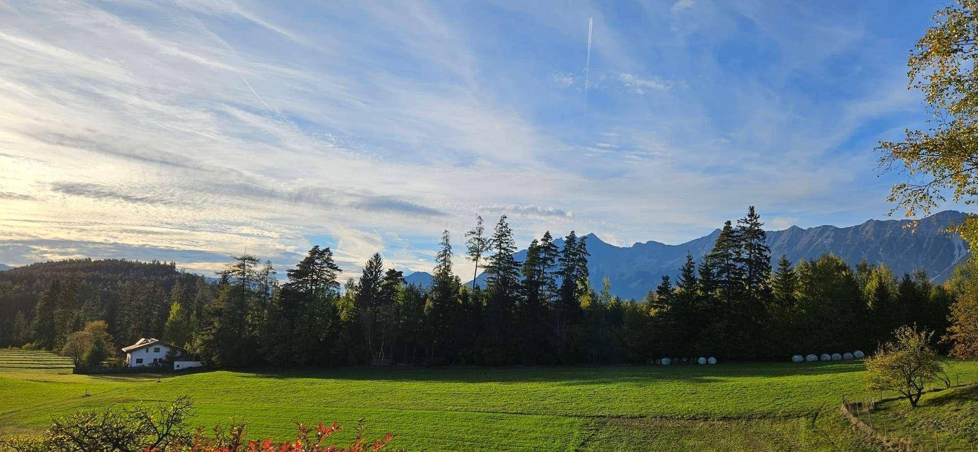 Weitläufige grüne Landschaft mit Wäldern, einem Haus und majestätischen Bergen im Hintergrund unter blauem Himmel.