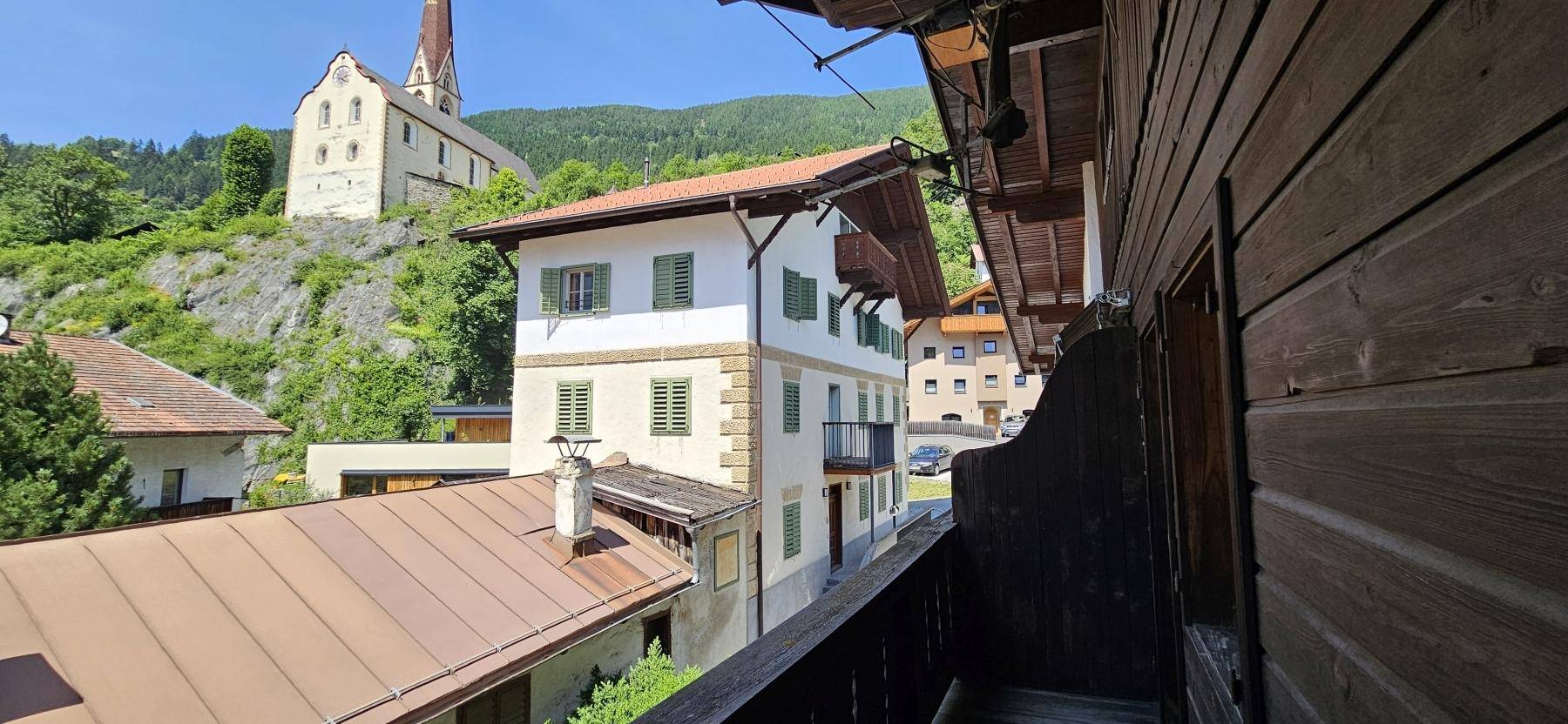 Blick vom Holzbalkon auf die umliegende Berglandschaft und ein traditionelles Dorf mit Kirche.