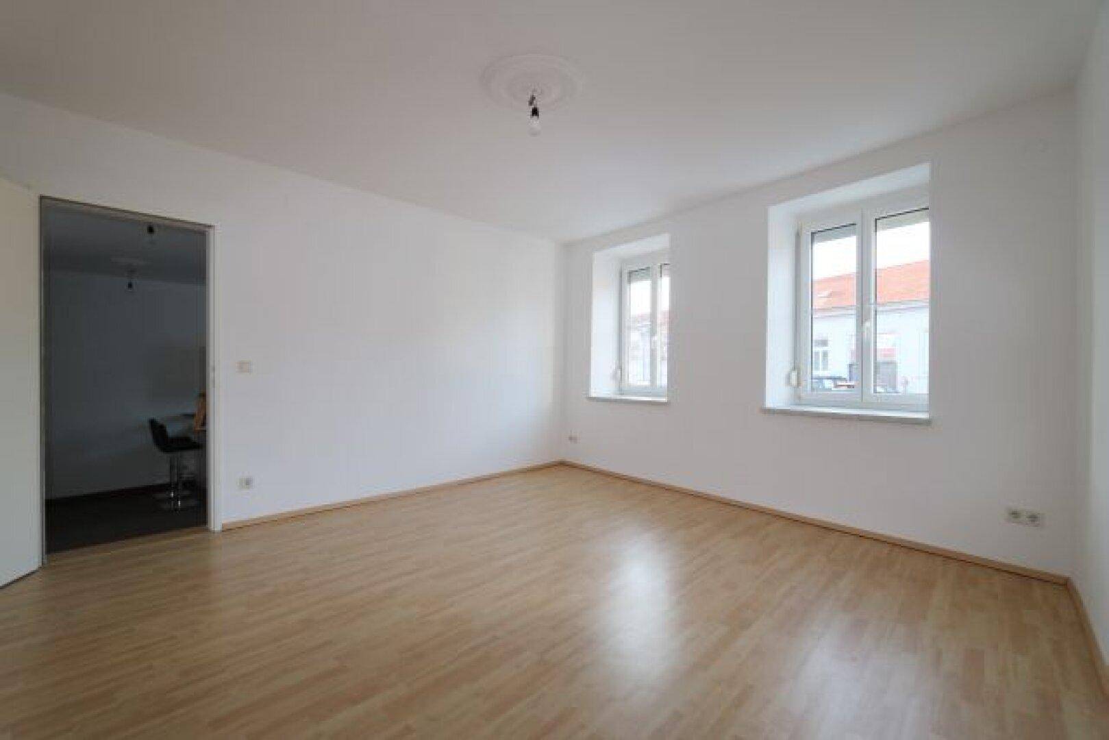 Geräumiges Wohnzimmer mit hellem Laminatboden und zwei großen Fenstern.