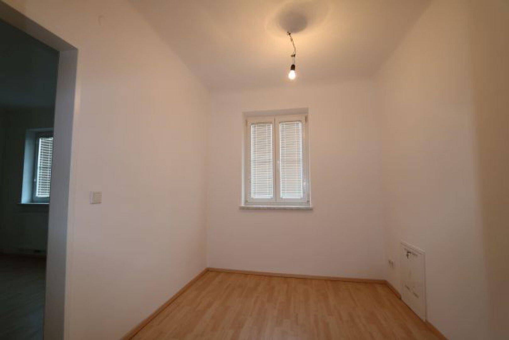 Helles Zimmer mit Laminatboden und einem Fenster mit Jalousien.