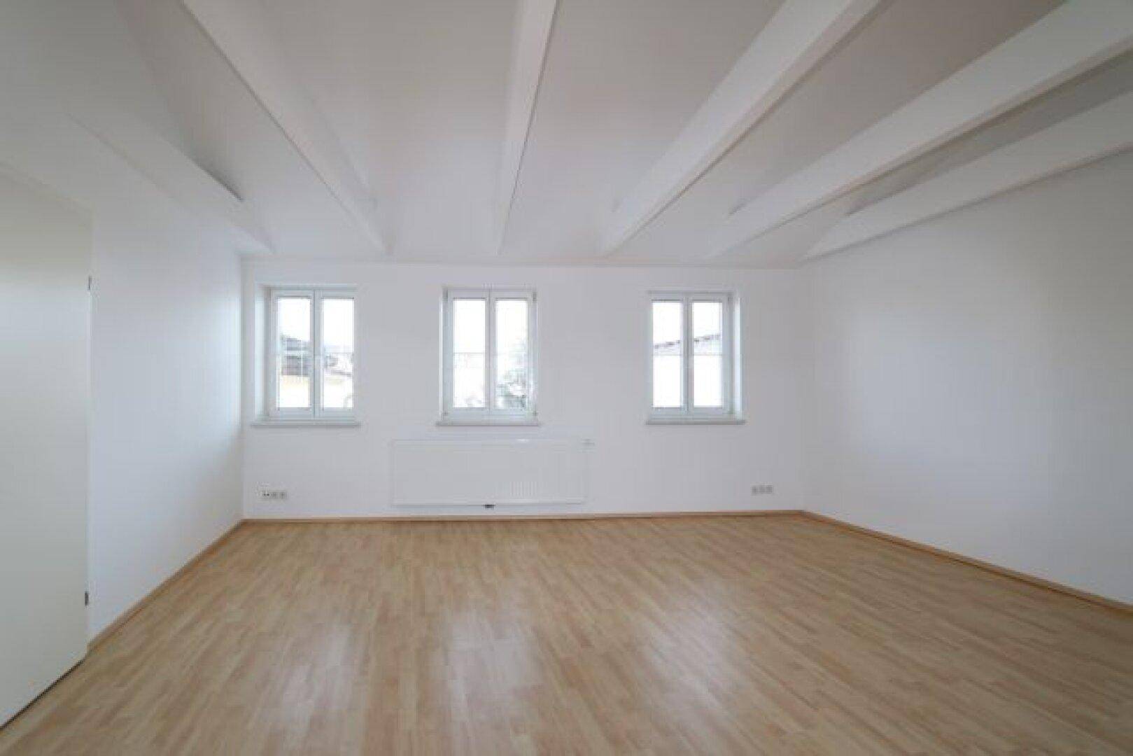 Geräumiges Dachgeschoss mit drei Fenstern, sichtbaren Balken und Laminatboden.