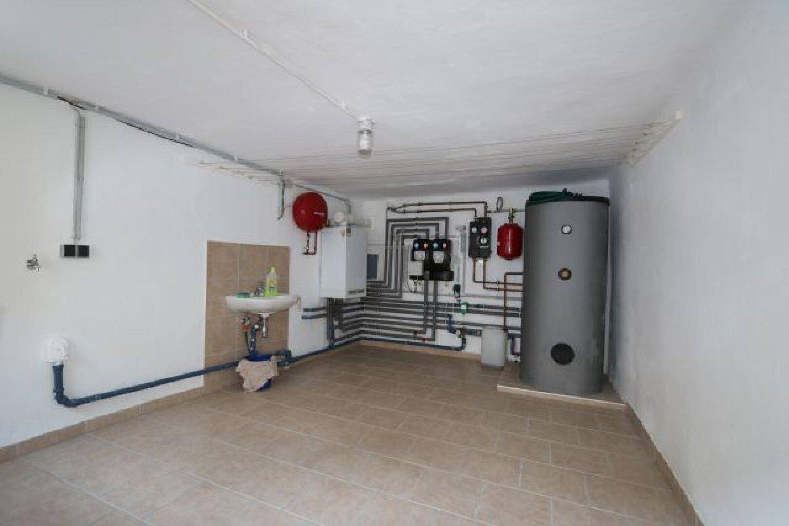 Technikraum im Keller mit Warmwasserspeicher, Rohrleitungen und Waschbecken.