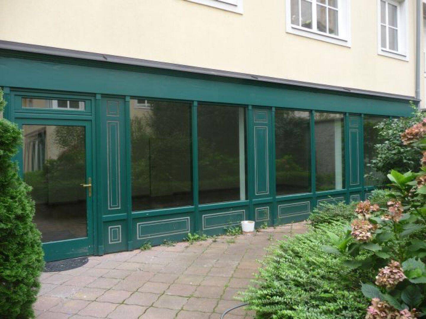 Grüne Ladenfront mit großen Schaufenstern und Eingangstür, umgeben von Sträuchern und einem gepflasterten Bereich.
