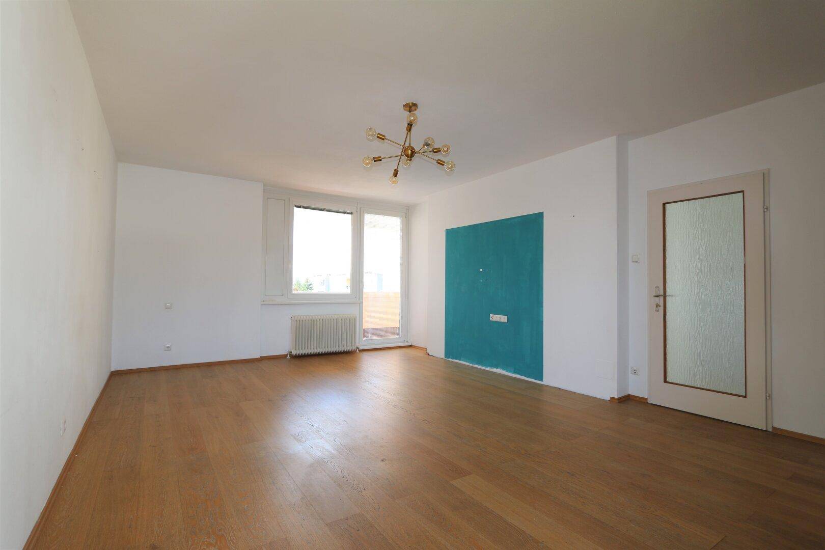 Helles, leeres Wohnzimmer mit Holzboden, weiß gestrichenen Wänden und einem blauen Akzentbereich.