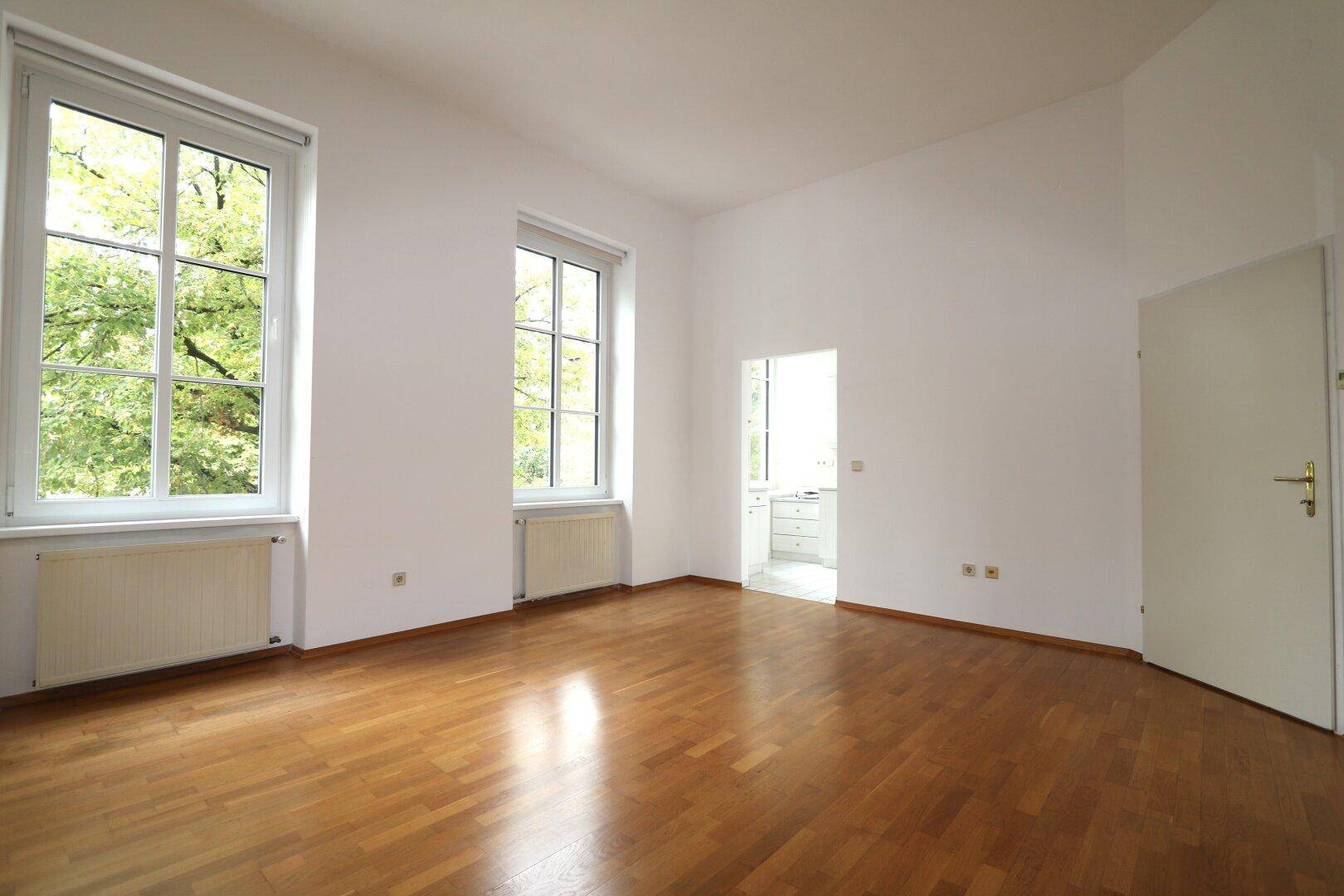Helles Zimmer mit Parkettboden, hohen Fenstern und Blick ins Grüne, ideal als Wohnzimmer nutzbar.