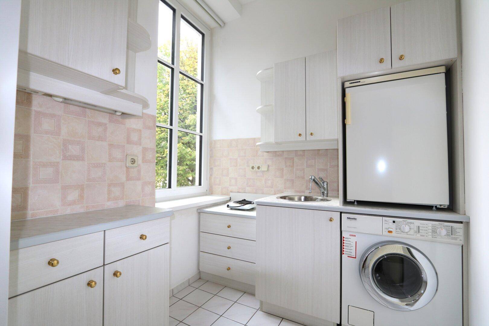 Kleine Küche mit weißen Schränken, Fliesenboden und Fenster, ausgestattet mit Waschmaschine und Kühlschrank.