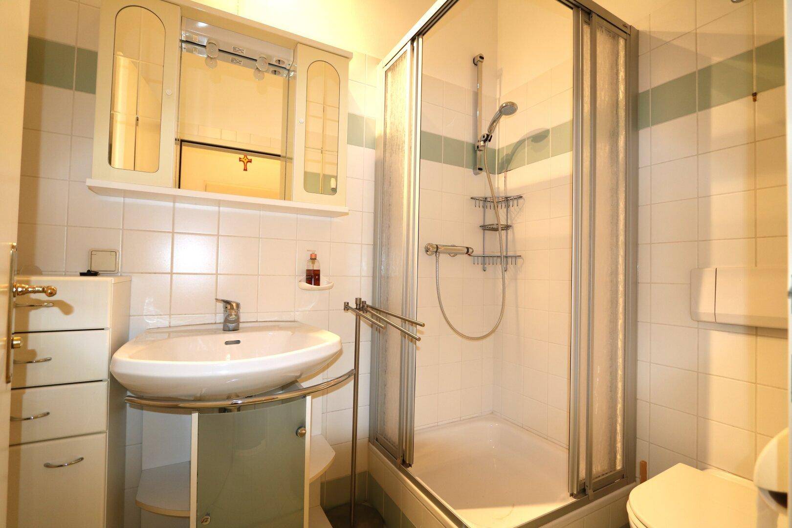 Badezimmer mit Duschkabine, Waschbecken und Spiegelschrank, ausgestattet mit weißen Fliesen und grünen Akzenten.