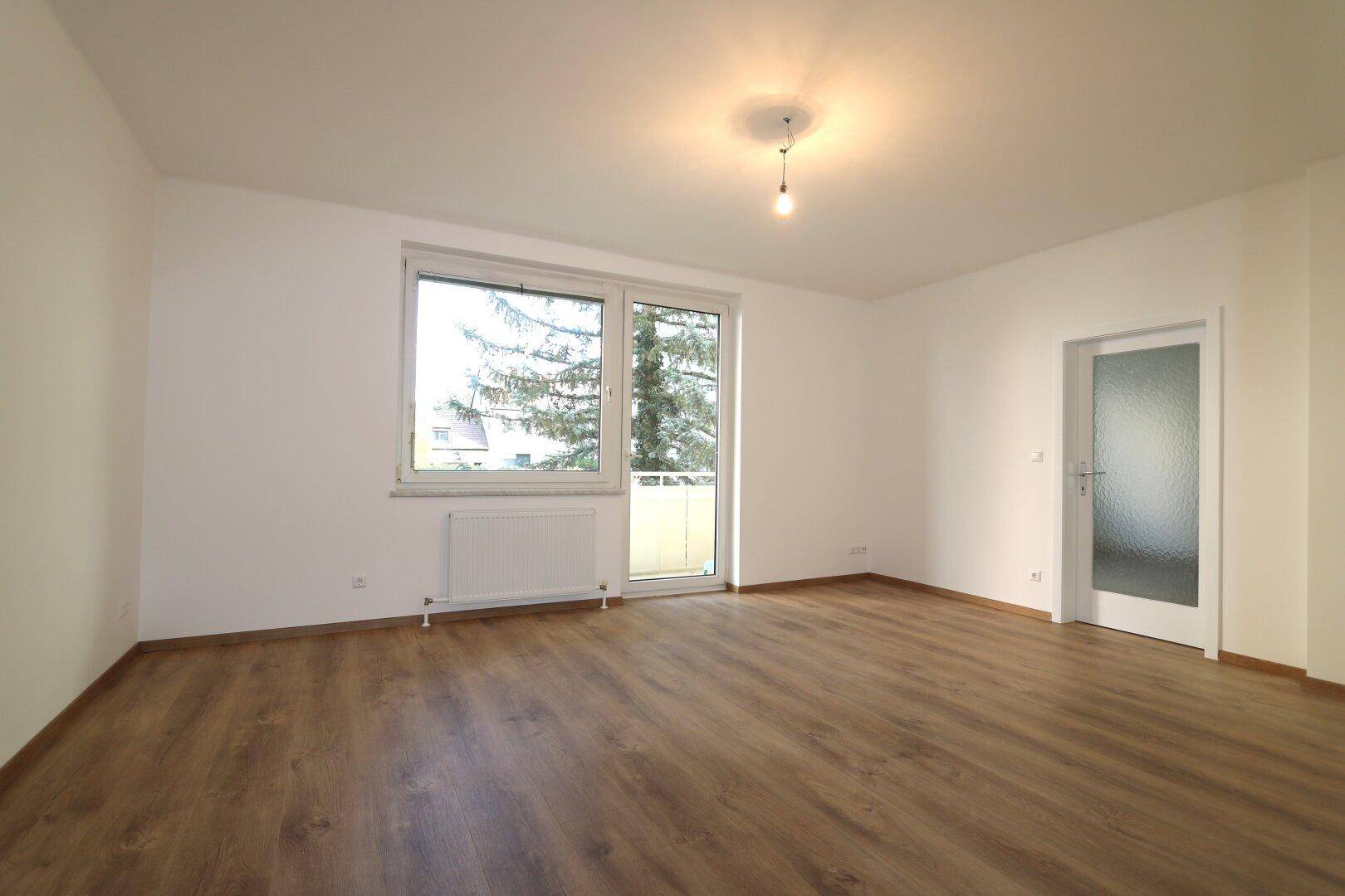 Heller Wohnraum mit Laminatboden, weißen Wänden und Zugang zu einem Balkon.