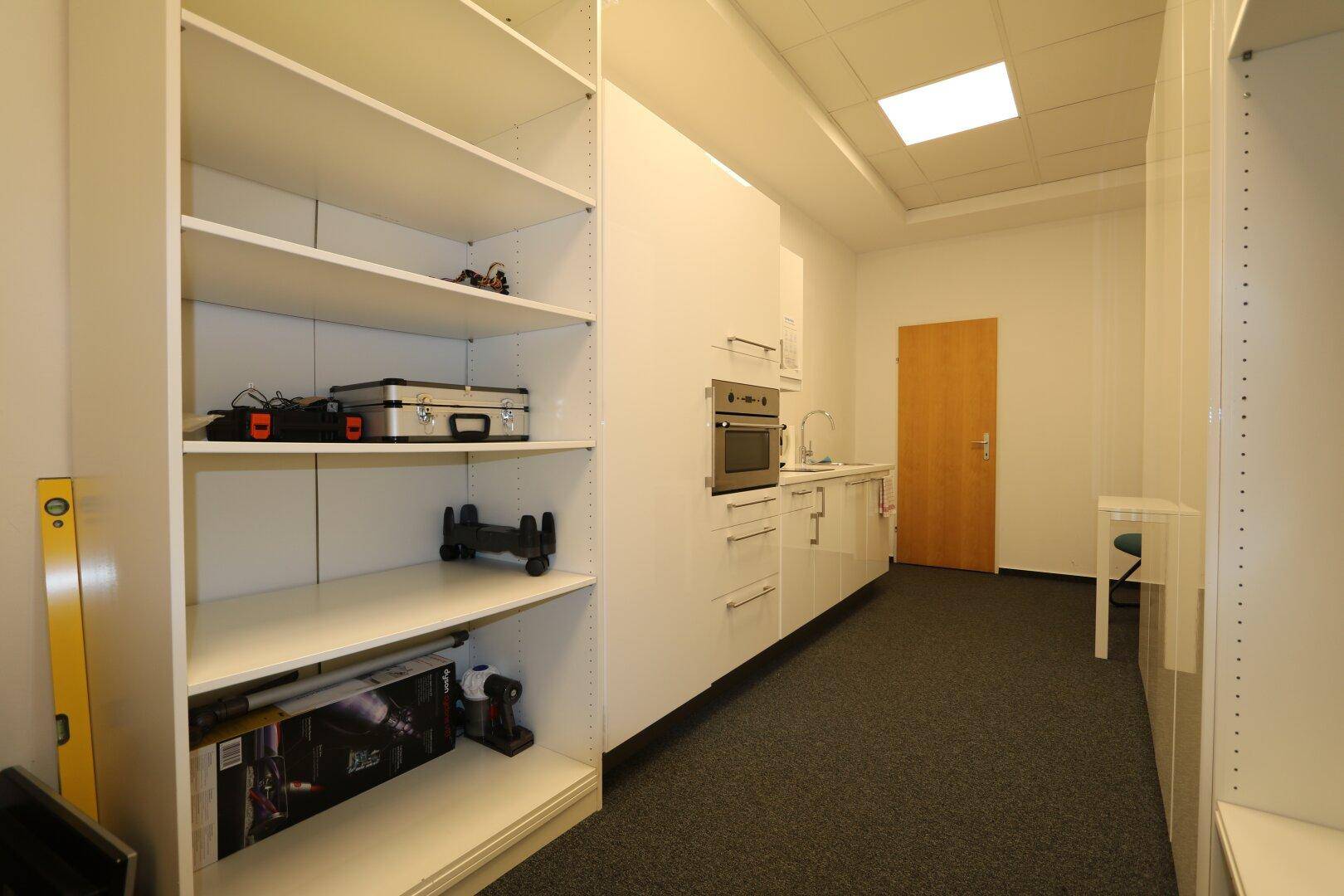 Kleine Teeküche mit weißen Schränken, Spüle und integriertem Ofen, neben Regalen für Lagerung.