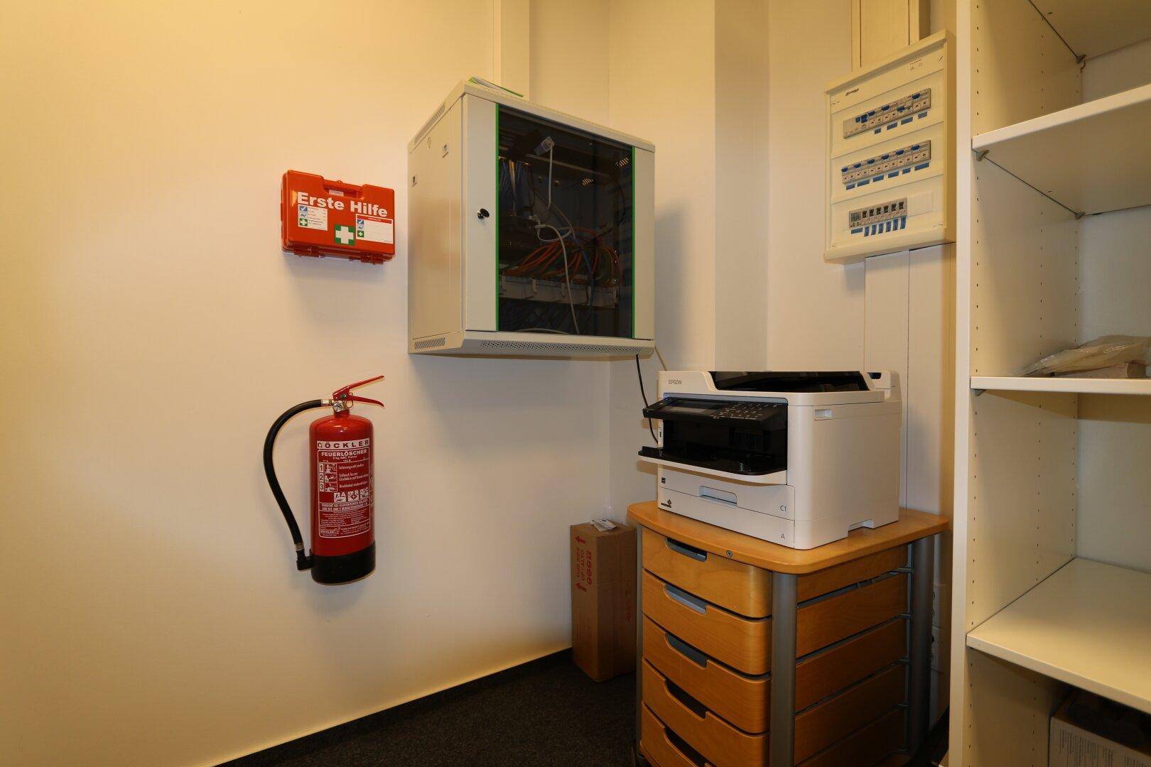 Funktionaler Technikraum mit Serverschrank, Drucker, Feuerlöscher und Erste-Hilfe-Kasten sowie Regalen und Elektroverteilung.