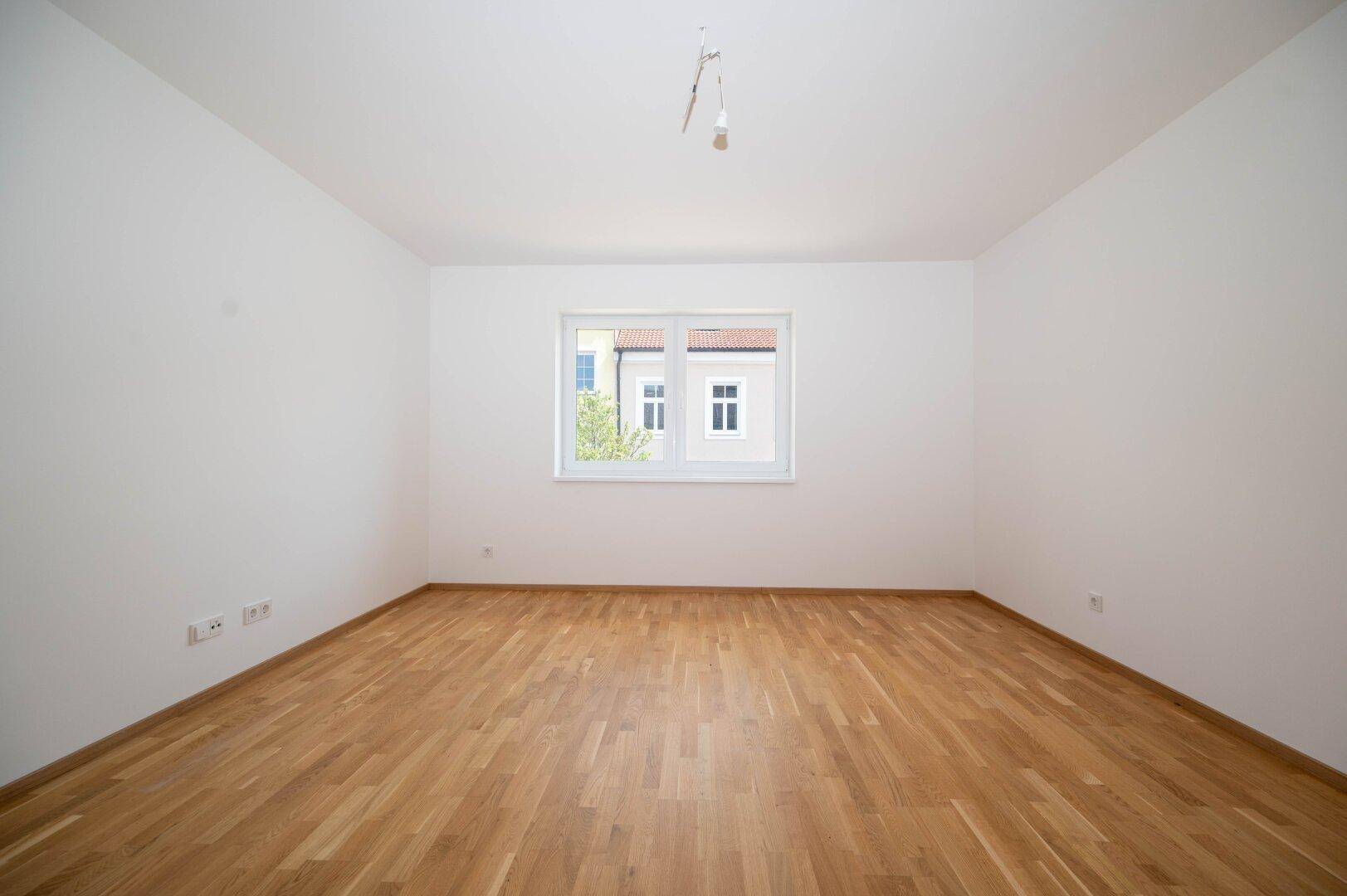 Unmöbliertes Zimmer mit Parkettboden und einem Fenster, das den Blick auf die Umgebung freigibt.