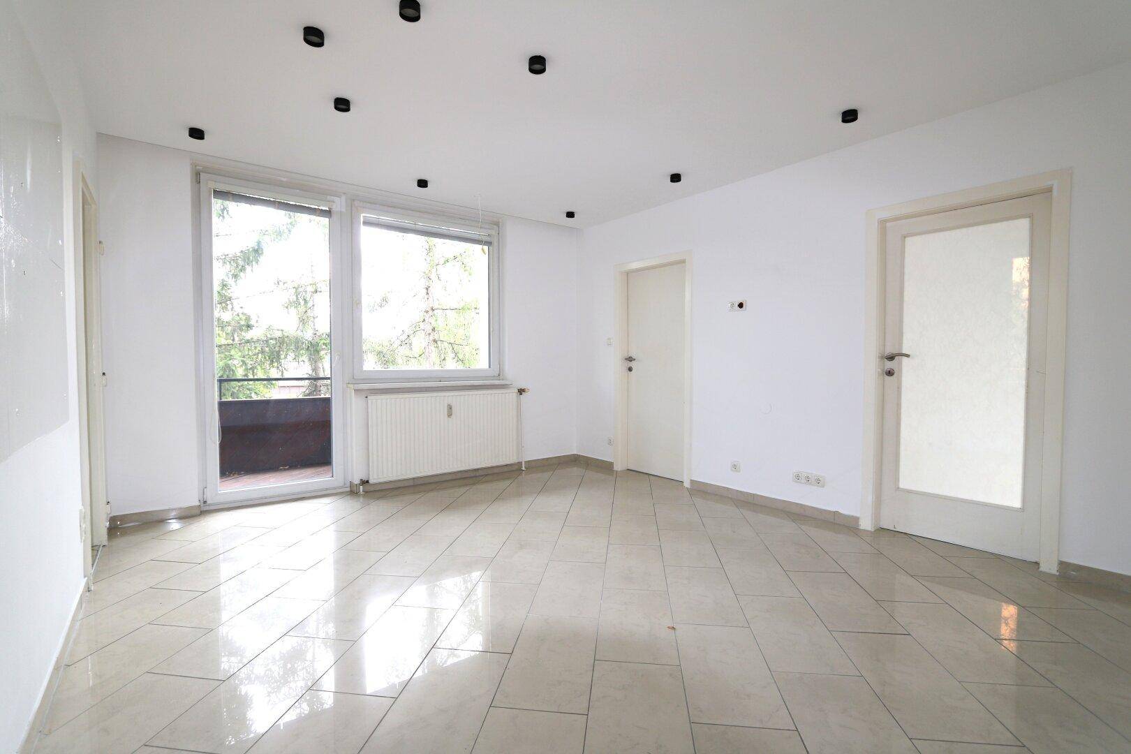 Helles Wohnzimmer mit Fliesenboden, Zugang zum Balkon und Blick ins Grüne.