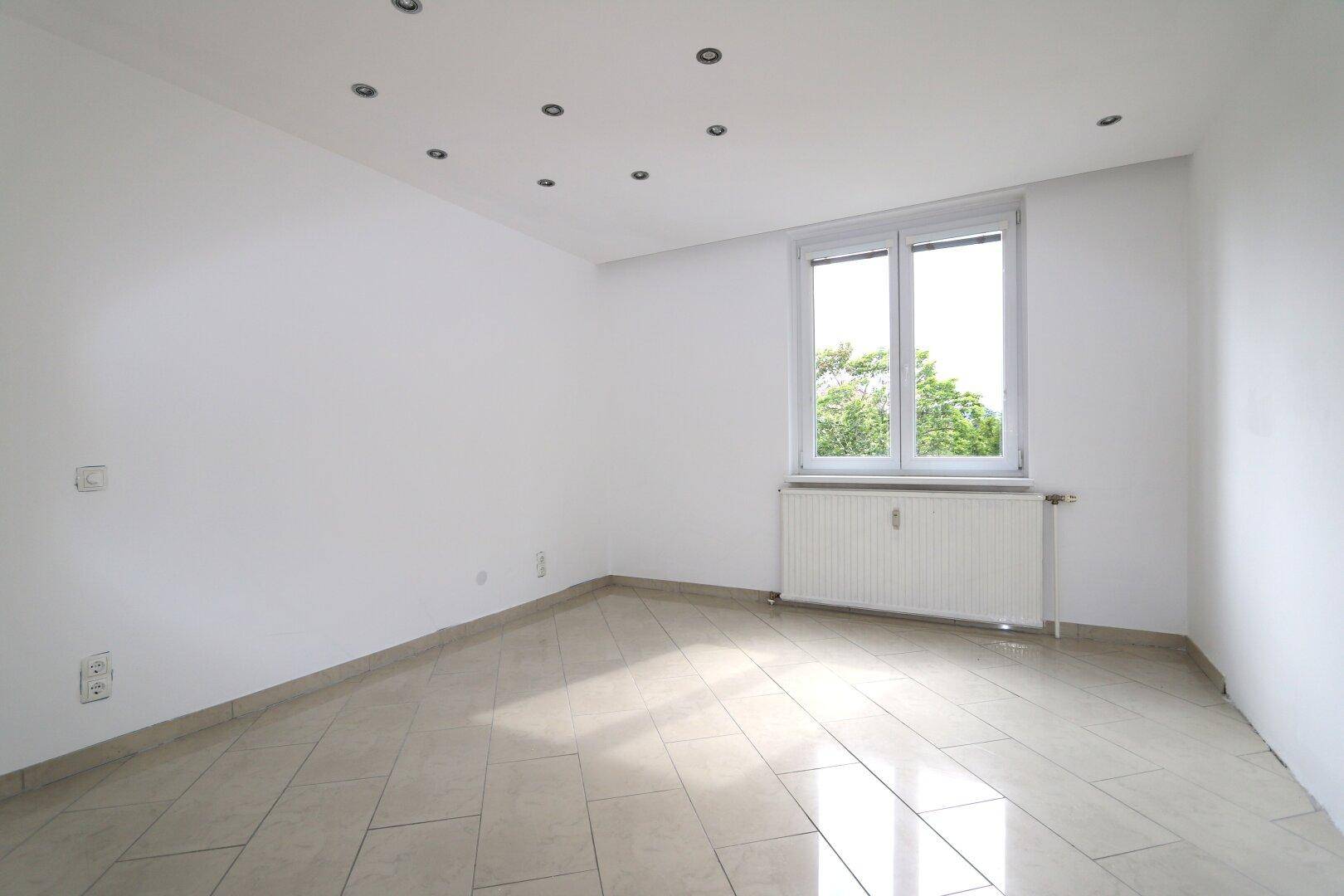 Helles Schlafzimmer mit Fliesenboden, Deckenstrahlern und Fenster mit Blick ins Grüne.