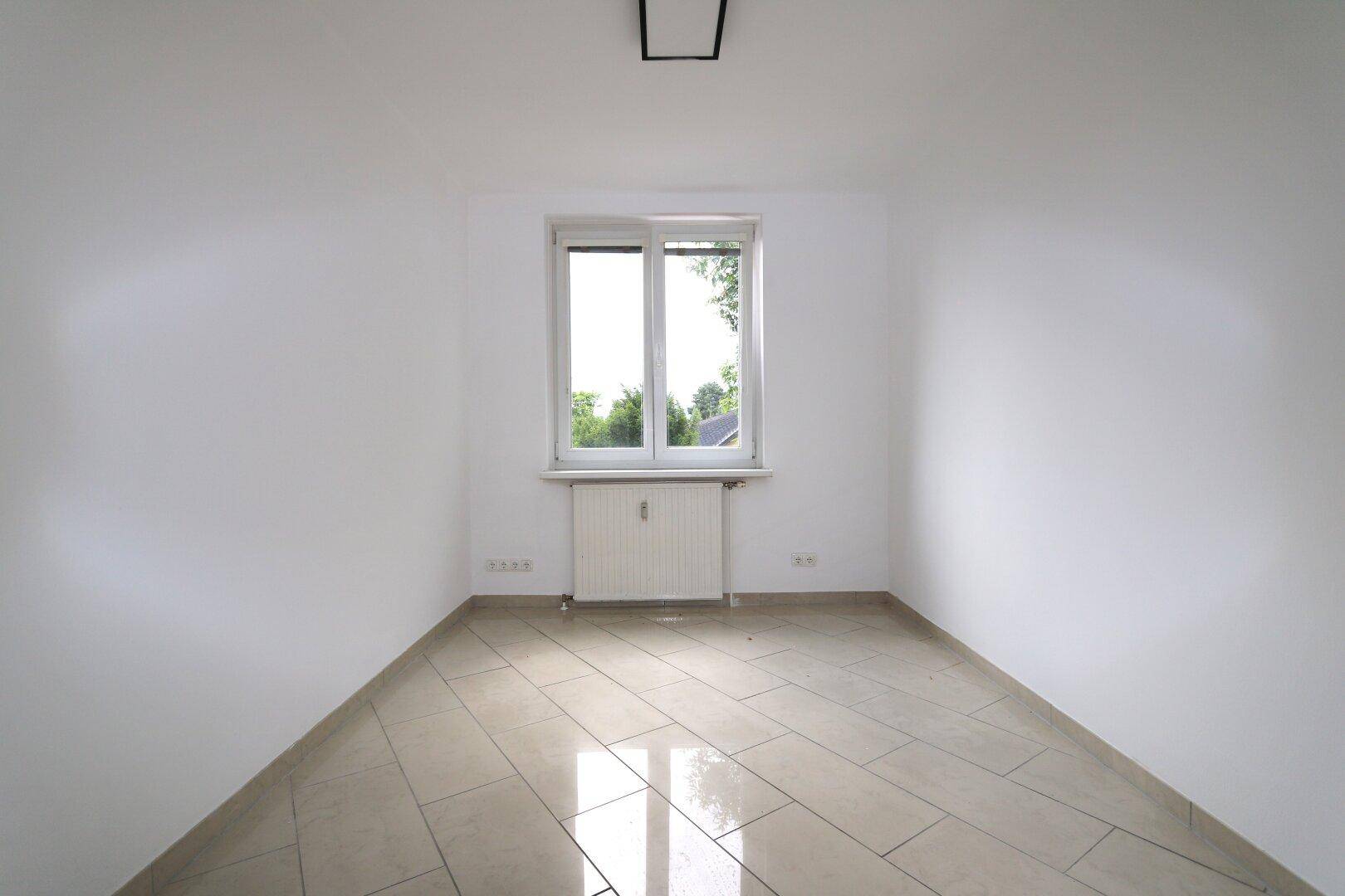 Freundliches Zimmer mit Fliesenboden, weiß gestrichenen Wänden und großem Fenster.