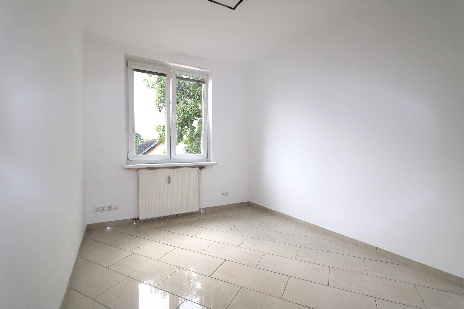 Helles Zimmer mit Fliesenboden, weiß gestrichenen Wänden und Fenster mit Blick nach draußen.