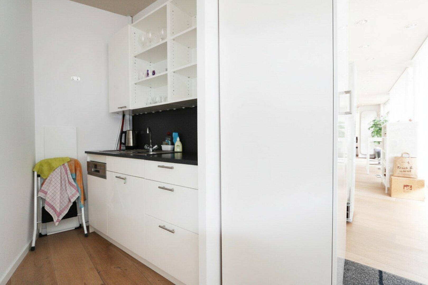 Kleine, moderne Küchenzeile mit weißen Schränken, schwarzer Arbeitsplatte und offenem Regal, inklusive Geschirrspüler.