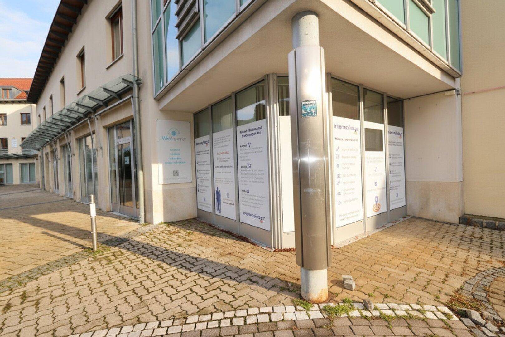 Außenansicht eines modernen Geschäftsgebäudes mit großen Schaufenstern und gepflastertem Vorplatz, der zum Eingang führt.
