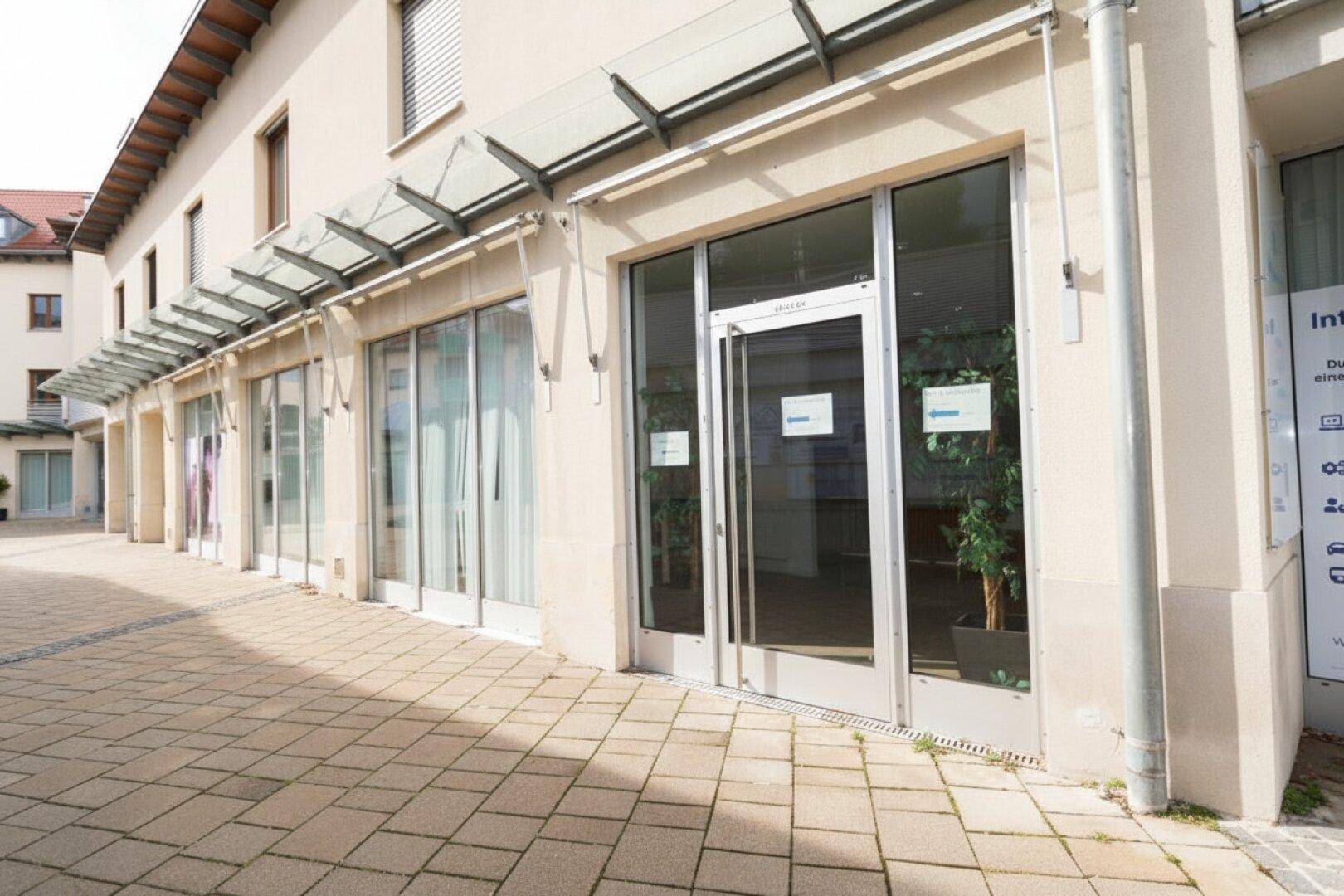 Eingangsbereich eines Geschäftsgebäudes mit großen Glasfenstern und einem Vordach aus Glas, das Schutz bietet.