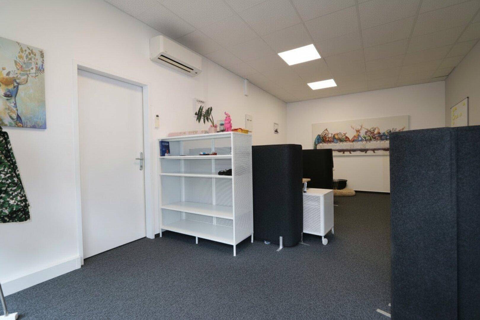 Büroansicht mit weißer Tür, offenem Regal und Klimaanlage an der Wand, funktional eingerichtet.