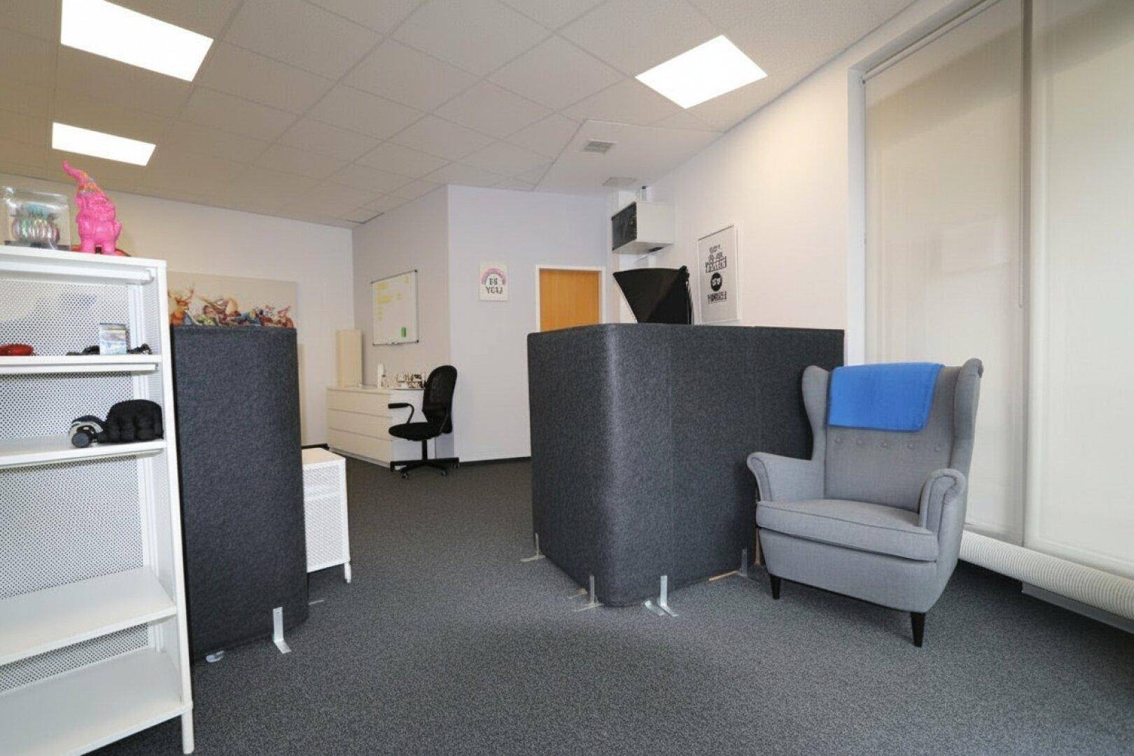 Büro mit gemütlichem Sessel, offenem Regal und weiteren Arbeitsplätzen im Hintergrund, einladend gestaltet.