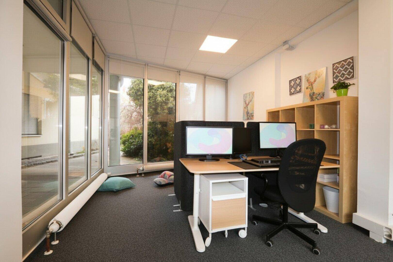 Helles Büro mit großen Fenstern, die einen Blick auf die begrünte Außenanlage bieten, viel Tageslicht.