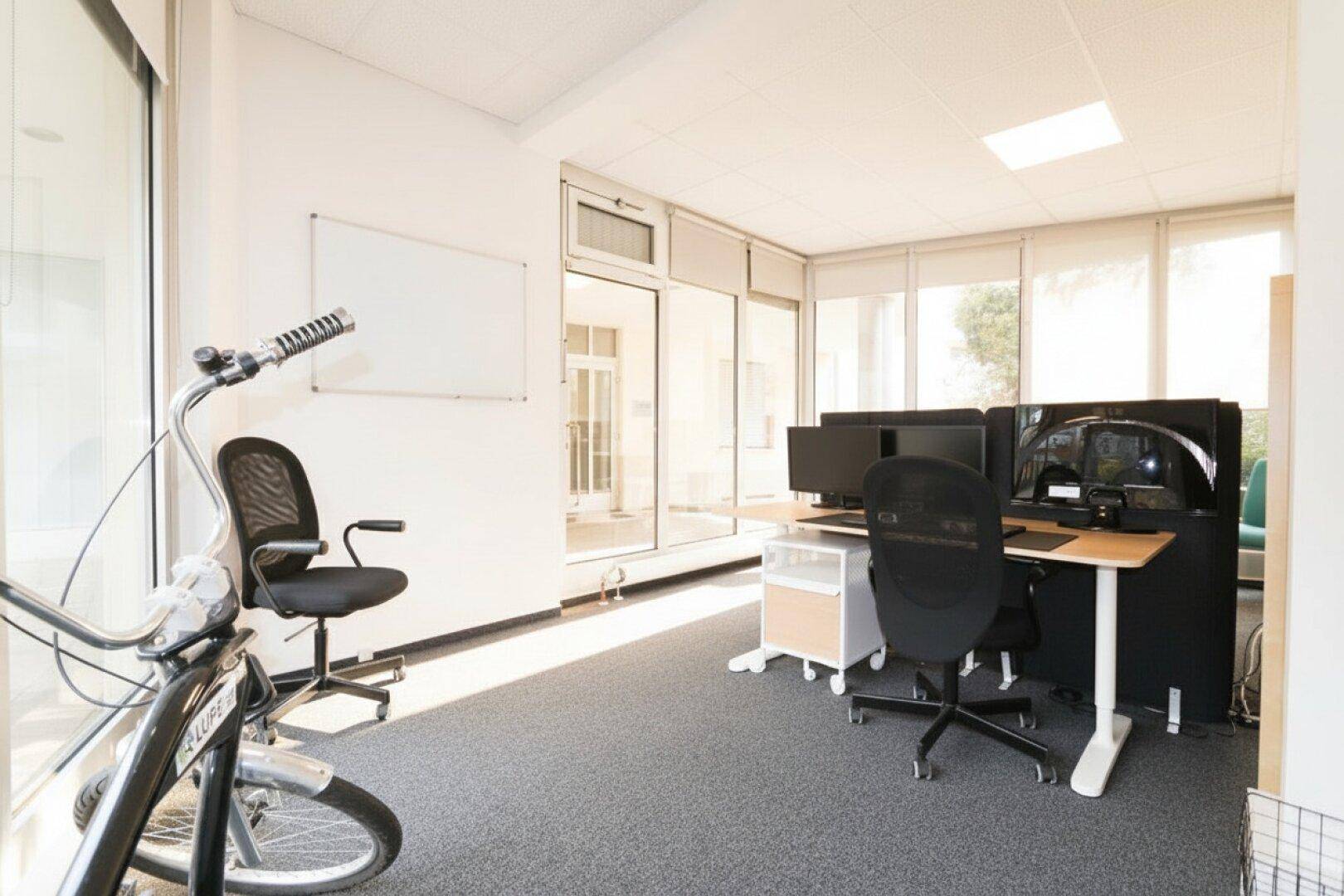 Geräumiges Büro mit viel Tageslicht durch große Fenster und einem Fahrrad als Dekoration, offene Gestaltung.