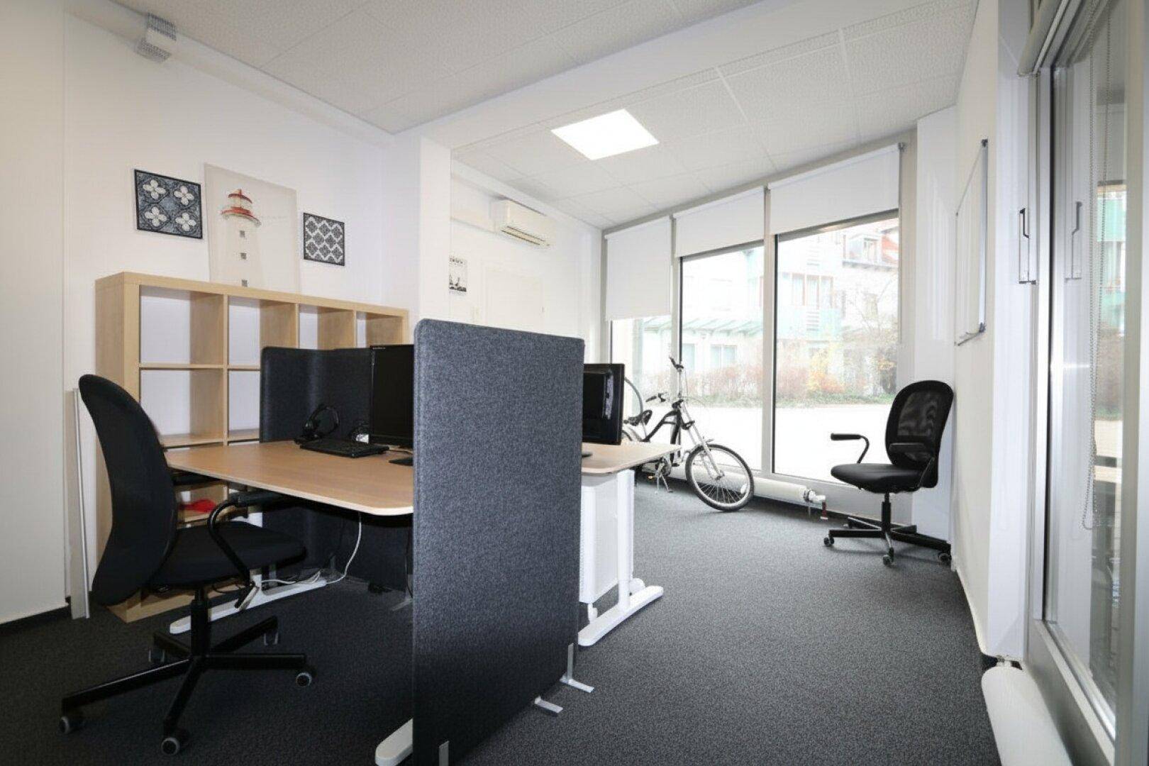 Offener Bürobereich mit mehreren Arbeitsplätzen, Raumteilern und großen Fenstern zur Straße, modern eingerichtet.