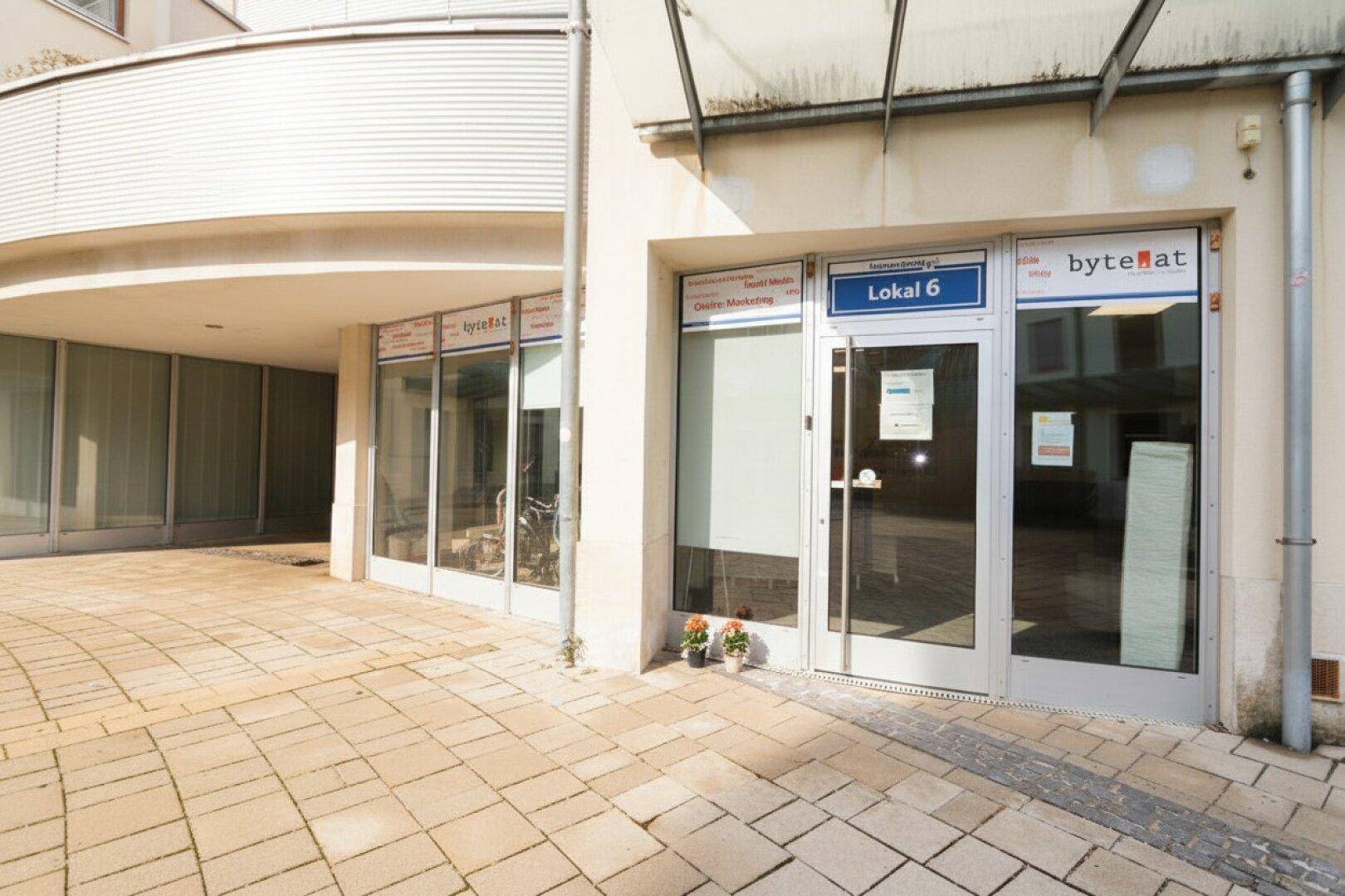 Eingangsbereich eines Geschäfts mit großen Fenstern und gepflastertem Vorplatz, einladende Fassade.