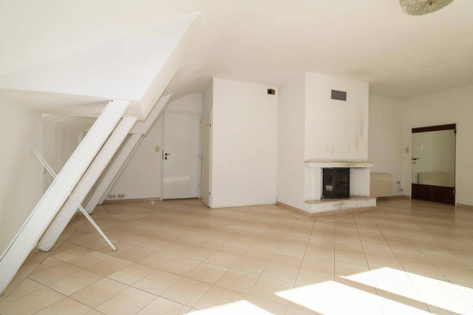 Geräumiges Wohnzimmer im Dachgeschoss mit Kamin und Fliesenboden, bietet viel Licht.