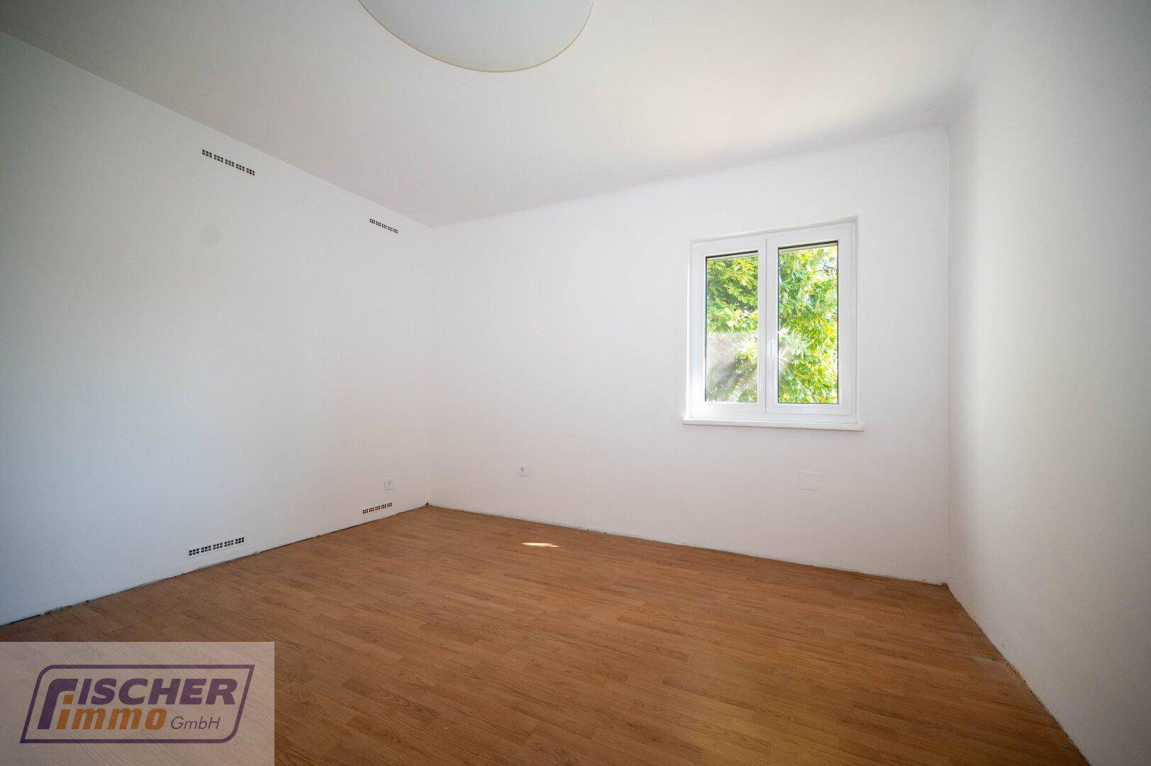 Leeres Zimmer mit hellem Laminatboden und einem Fenster mit Blick ins Grüne.