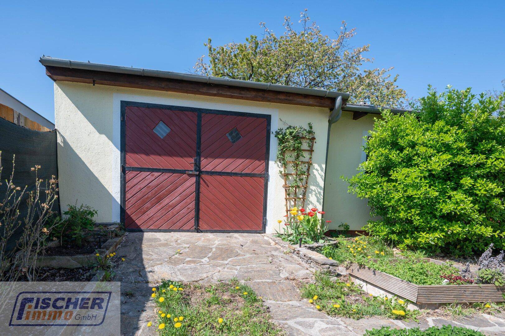 Externe Garage mit roten Holztoren und einem kleinen Vorgarten mit Blumen.