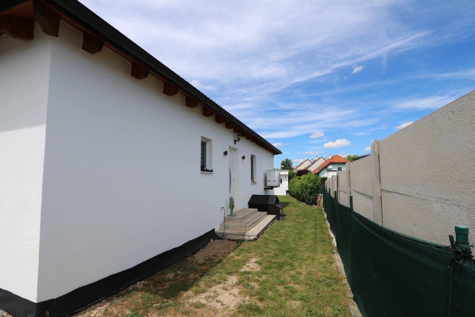 Weiße Hausfassade mit einem schmalen Grasweg und einem kleinen Treppenaufgang unter blauem Himmel.