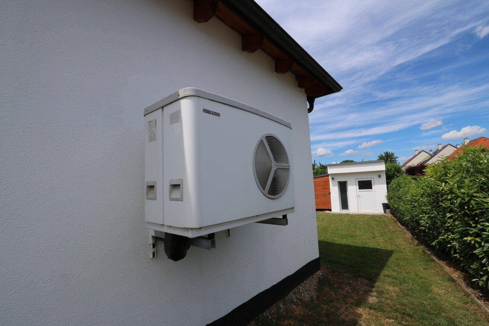 Eine Wärmepumpe ist an der weißen Außenwand des Hauses montiert, umgeben von grünen Büschen.